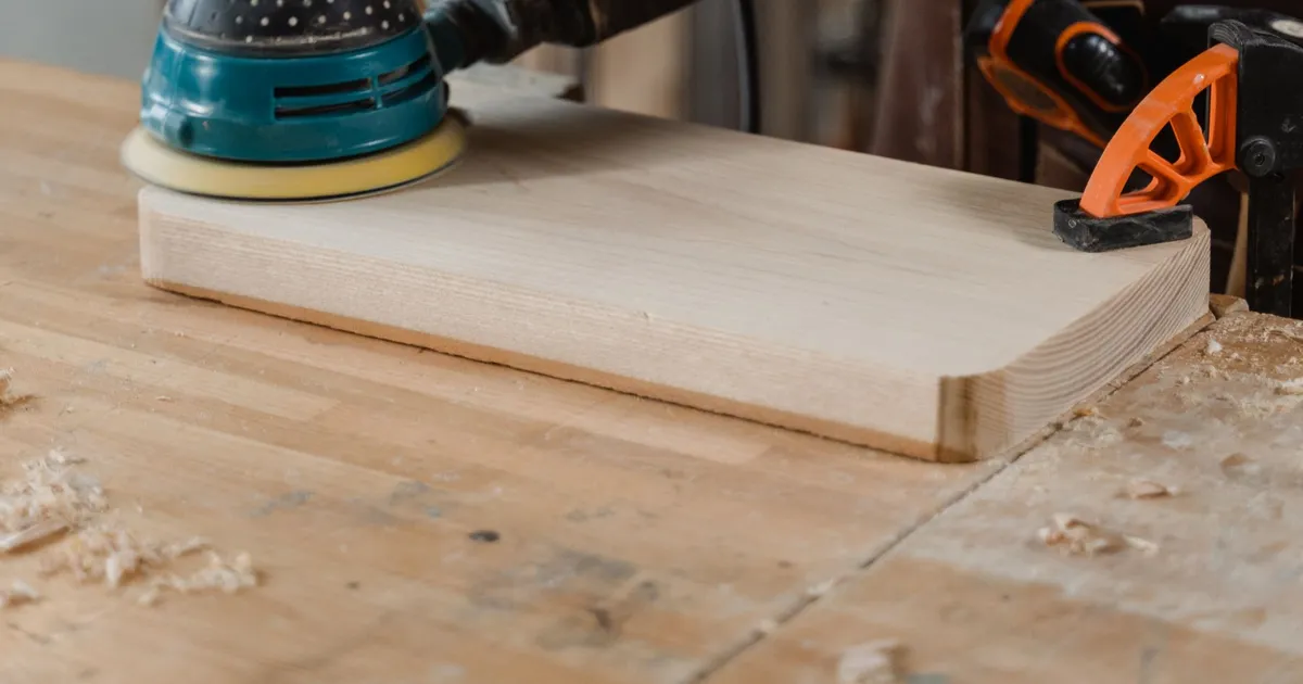 Orbital sander moving over a wooden board with a layered edge on a workbench, with wood dust around it